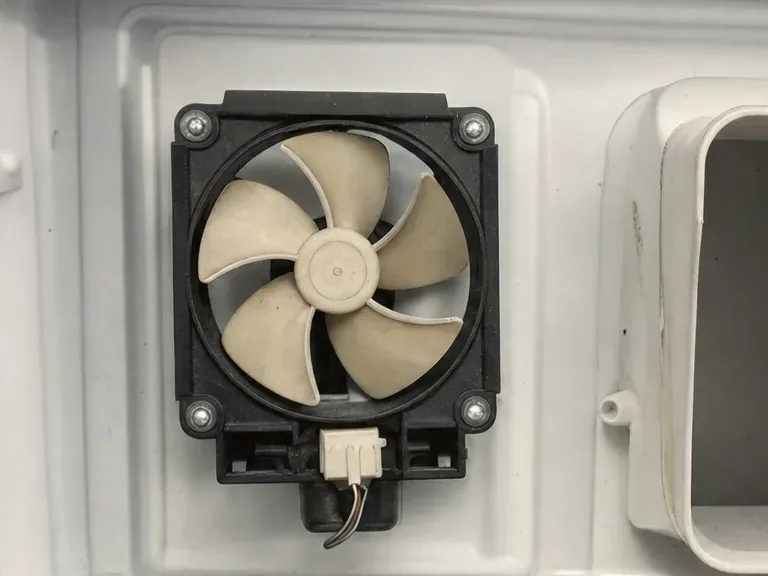 Square black-framed fan with beige blades mounted on the white back wall of a freezer compartment.