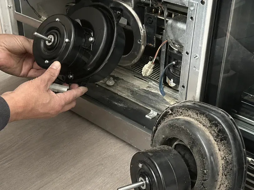 Technician holding a black fan motor at the open base of a refrigerator, a dust-coated old motor in the foreground.